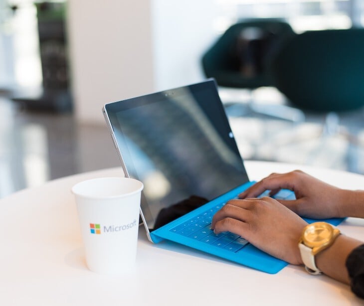 Image of someone typing on a computer. Next to them is a cup labelled with the Microsoft logo. 