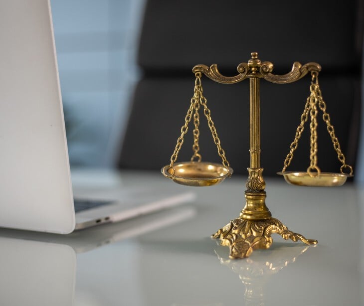 Image of the gold scales of justice on a table. In the background is a laptop and desk chair. 